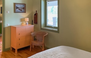 A bedroom with a wooden dresser, a wicker chair, a bed with a white duvet, a window with blinds, and a painting on the wall.