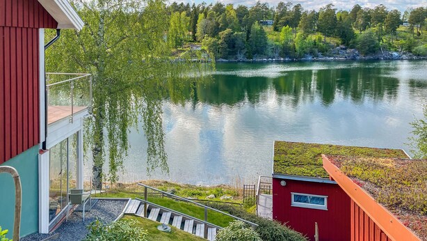 A lakeside view from a red building with a glass balcony overlooking a calm lake, surrounded by trees and a dock. - Pet friendly home in Vätö with sauna (Vätö)
