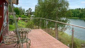 A wooden deck with a glass balustrade overlooking a lake, a metal table and chairs, and a red building in the background. - Pet friendly home in Vätö with sauna (Vätö)