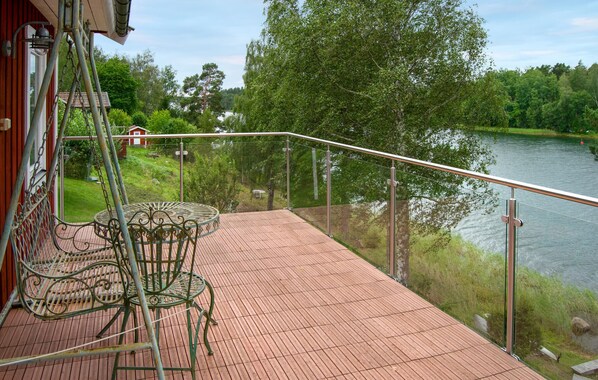 A wooden deck with a glass balustrade overlooking a lake, a metal table and chairs, and a red building in the background.