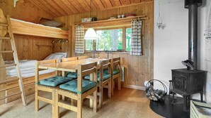 Een houten blokhut van binnen met stapelbedden, een eettafel met stoelen, een raam met gordijnen en een houtkachel.