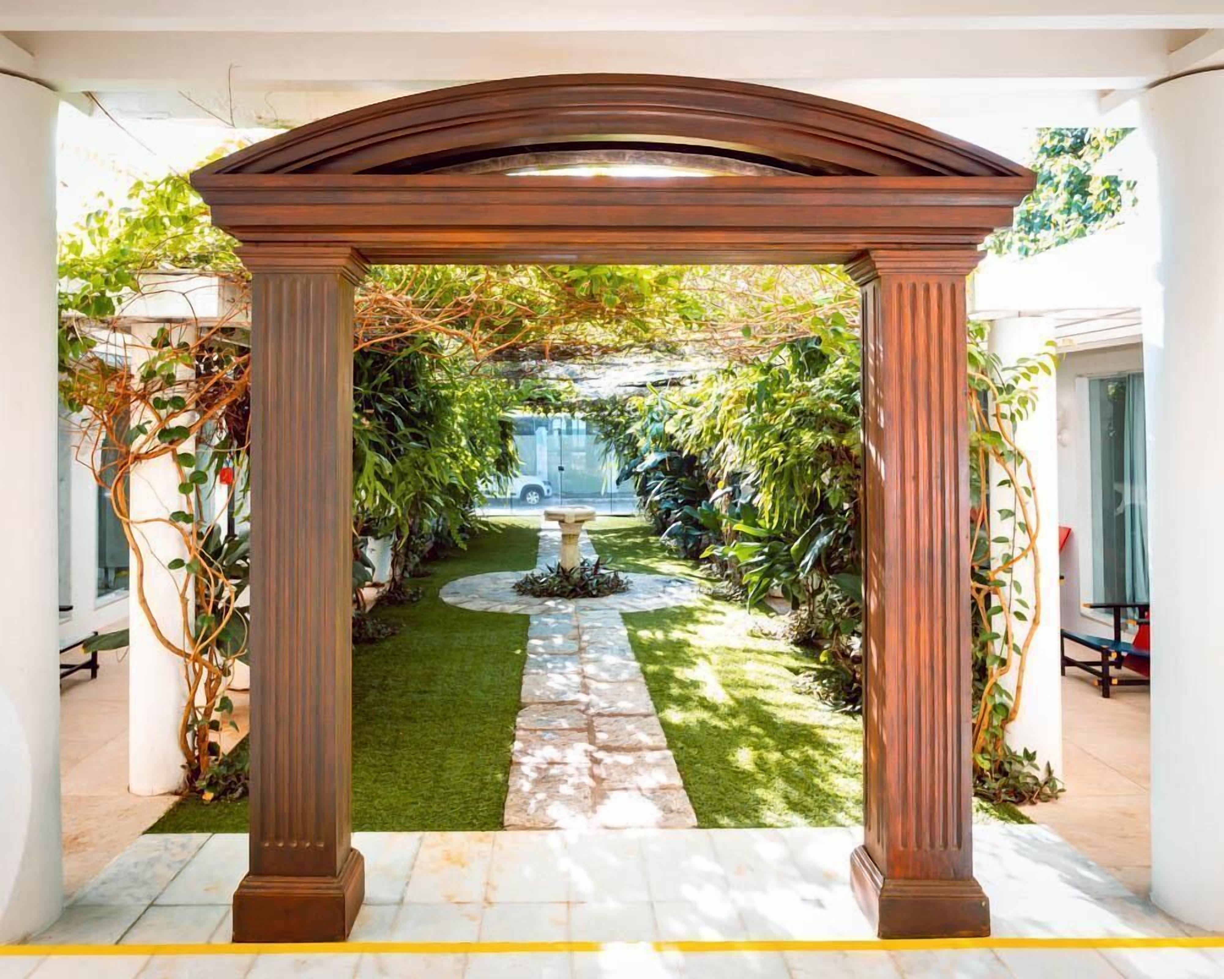 Un sendero cubierto en un jardín, con una fuente central rodeada de abundante vegetación y paredes blancas.