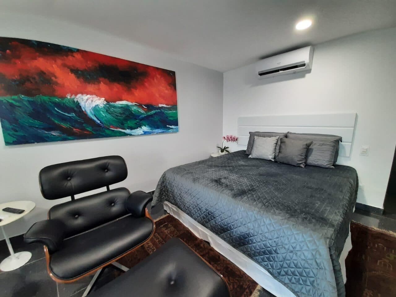 A bedroom with a bed, a headboard, a wall-mounted air conditioner, a painting, and a chair.