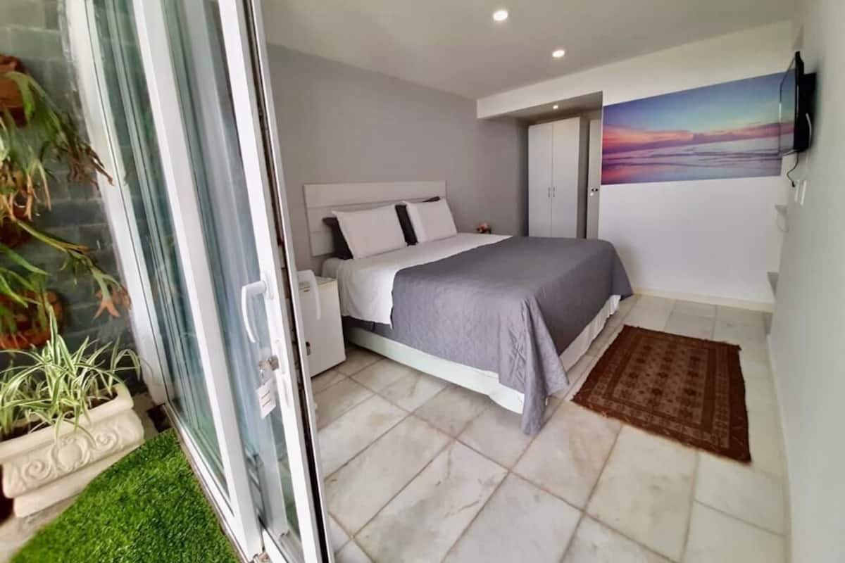 Un dormitorio con cama, una mesita de noche, televisor montado en la pared y una puerta que da a otra habitación.