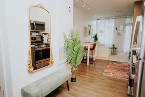 A modern living room with a large mirror, a bench, a dining area, and a kitchen with a microwave and stove.