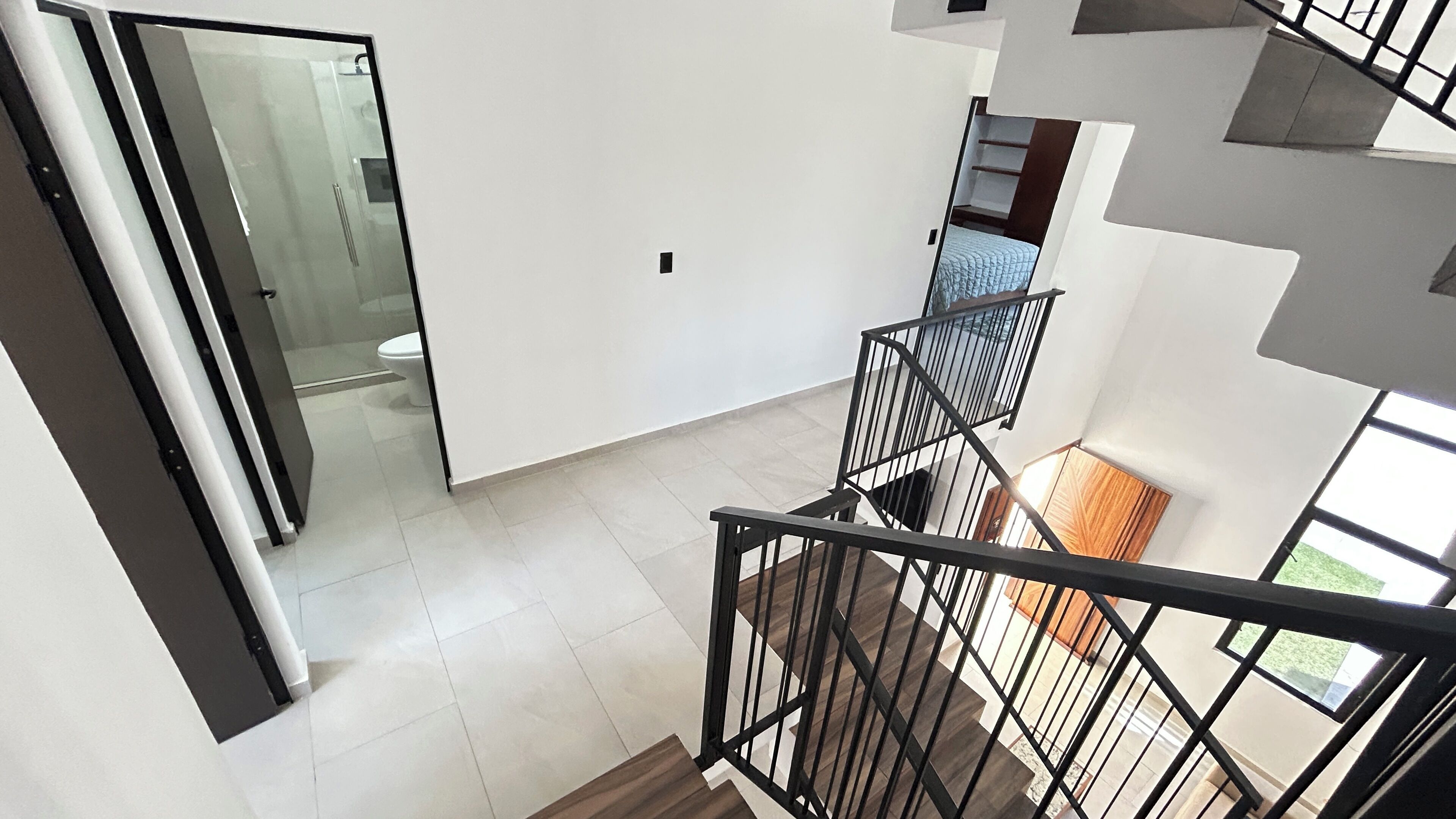 Un intérieur moderne avec un escalier, une porte vitrée donnant accès à une salle de bain et une vue sur un lit bleu.