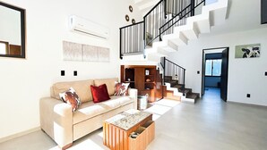 A spacious living room with a beige sofa, a coffee table, a staircase, and a modern air conditioning unit.