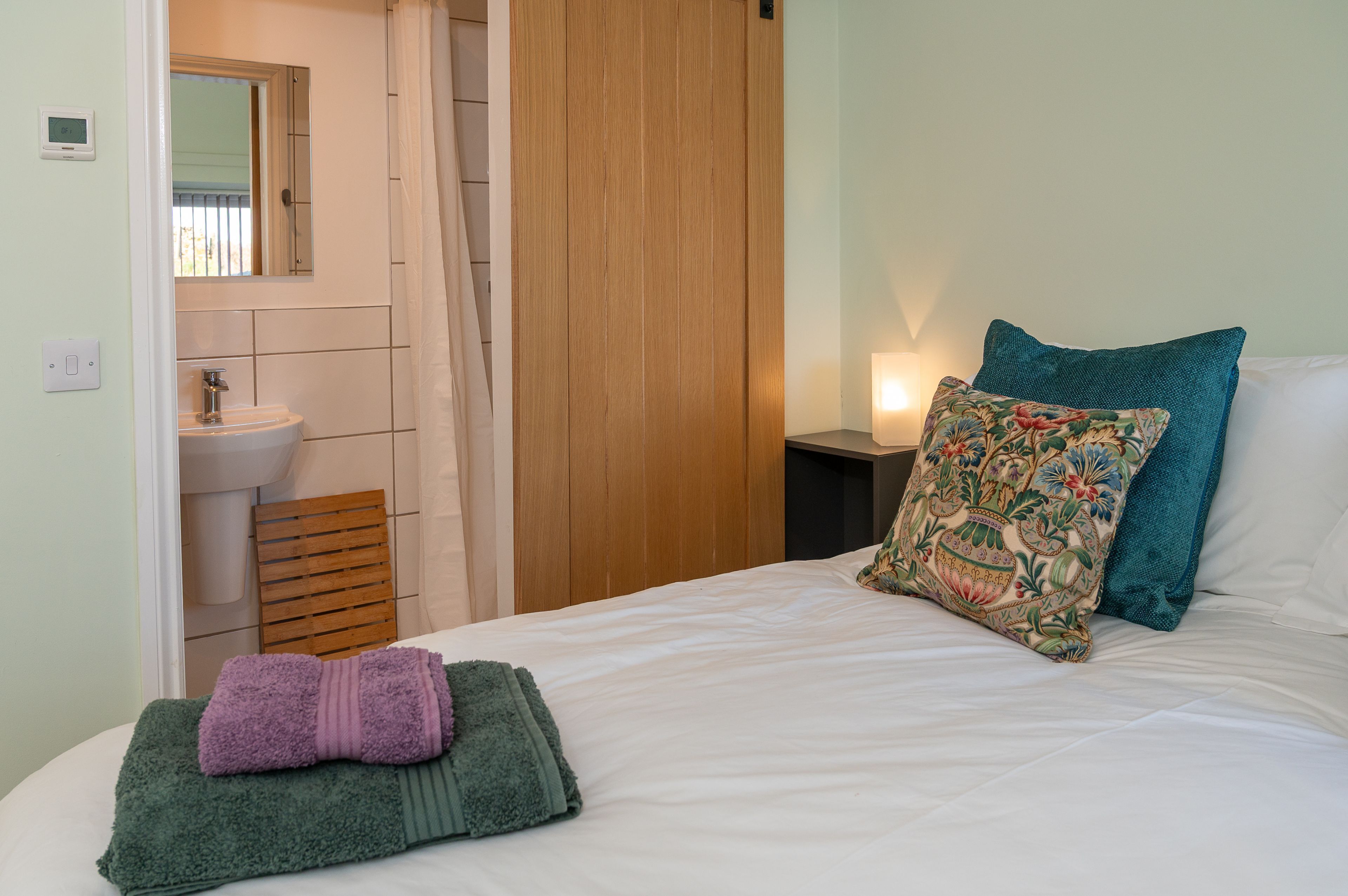Een slaapkamer met een bed, kussens, een nachtkastje met een lamp, een houten deur en een badkamer die zichtbaar is door een geopende deur.