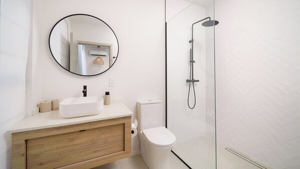 A modern bathroom with a white sink, round mirror, toilet, and glass-enclosed shower. - A Modern Vacation Villa - Sorrento 15 (Lower Carlton)