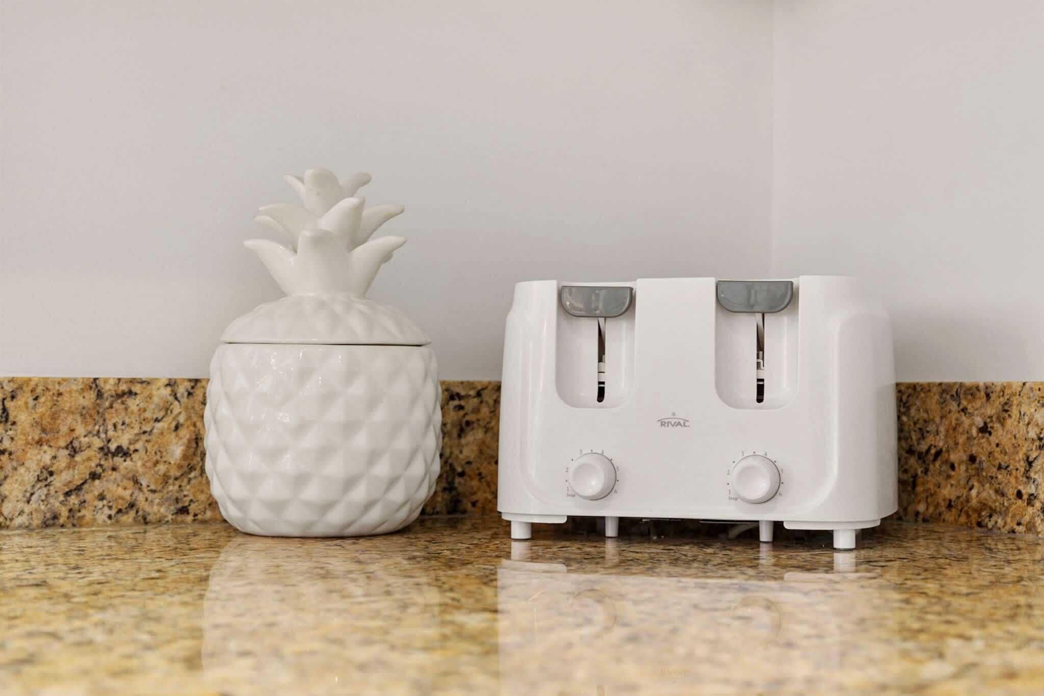 A white toaster and a white pineapple-shaped container on a granite countertop.