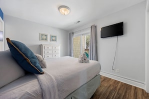 A bedroom with a bed, a TV mounted on the wall, a window with curtains, and a small white dresser.