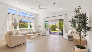 A spacious living room with a beige sofa set, a coffee table, and a side table. There is a potted plant and a large window with curtains. - Harlow House - Lounge poolside in Indian Wells (Indian Wells)