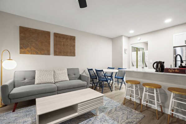 A modern living room with a grey sofa, blue chairs, a coffee bar, and a kitchen area with white cabinets and a stainless steel refrigerator.