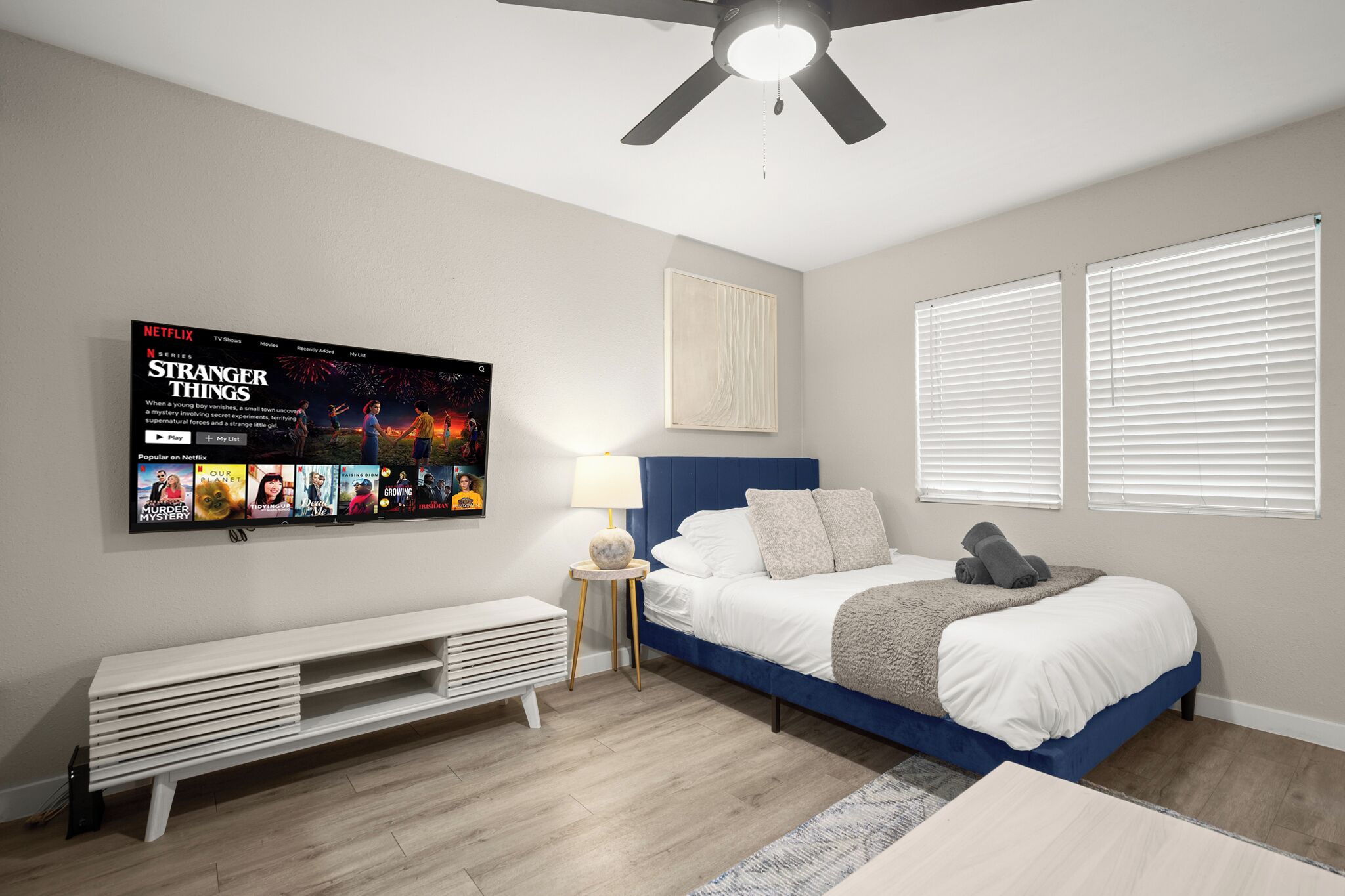 A modern bedroom with a bed, a TV mounted on the wall, a ceiling fan, and a window with blinds.