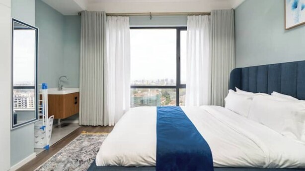 A hotel room with a large bed, a view of the city, a bathroom with a sink, and a window with curtains.