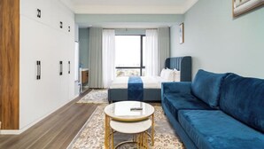 A modern hotel room with a blue sofa, a small round table, a bed with a blue blanket, and a large window with curtains.