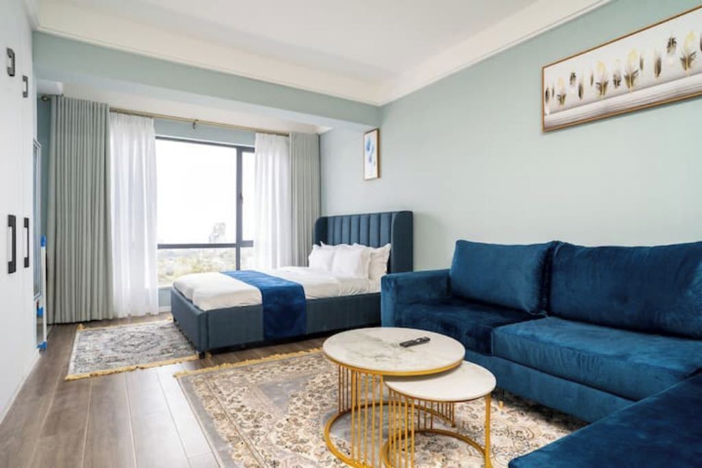 Et moderne hotelværelse med en seng, en blå sofa, et rundt sofabord og et stort vindue med gardiner.