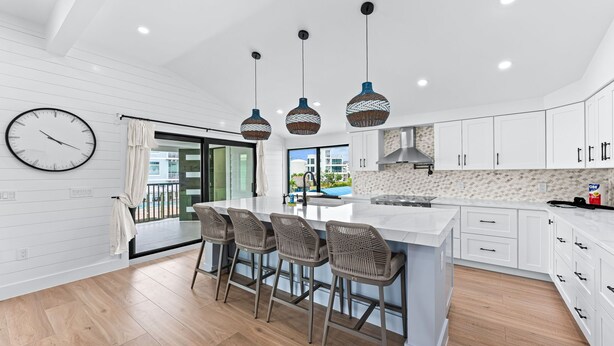Une cuisine moderne avec des meubles blancs, un grand îlot central doté de tabourets, des luminaires suspendus et une vue sur l’extérieur.