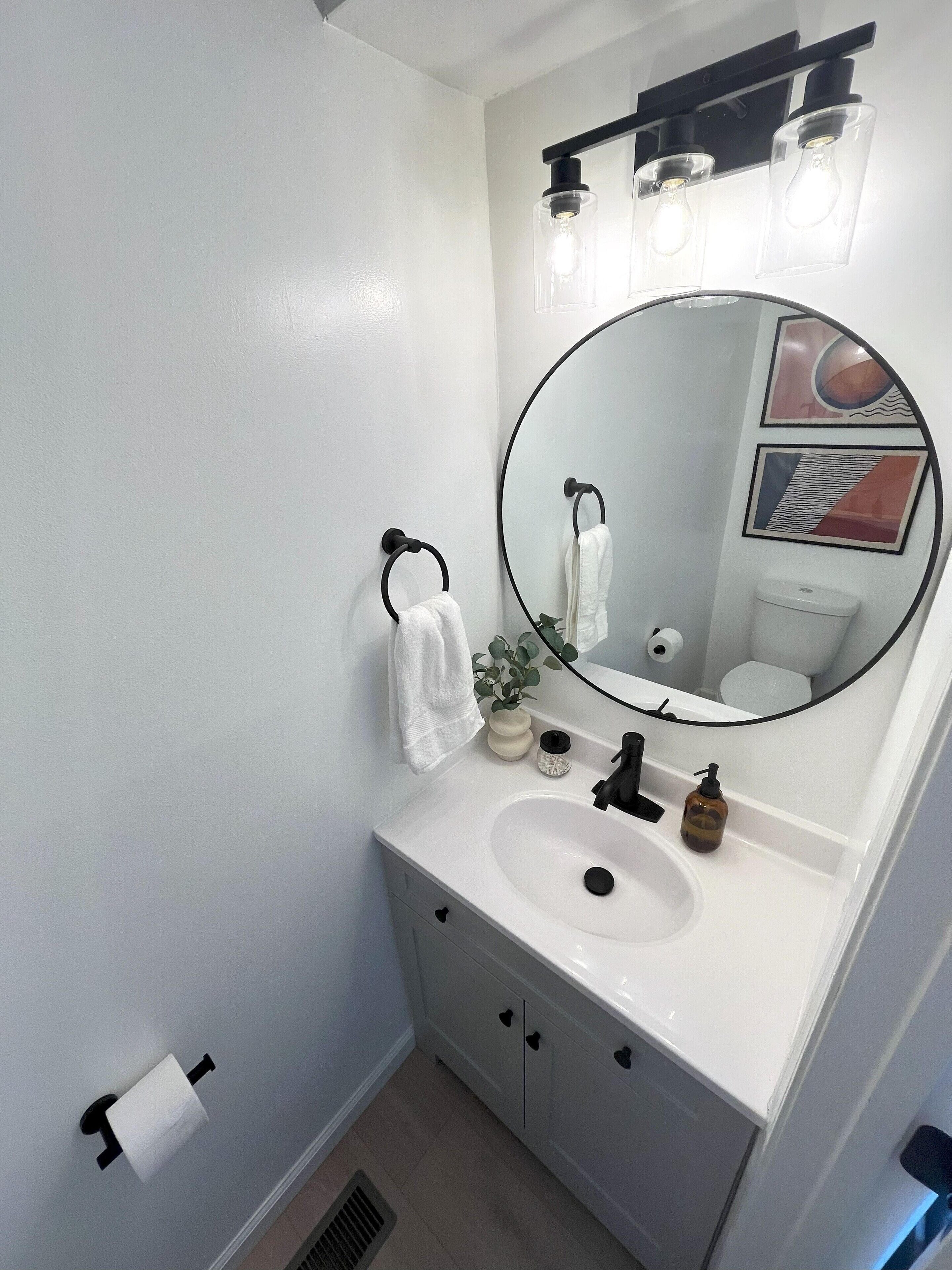 A bathroom with a white sink, black faucet, and a round mirror. There are two towel rings with white towels, a small plant, and a toilet paper holder on the wall.