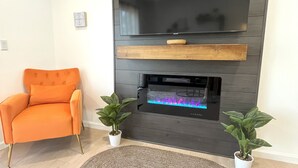 Uma sala de estar moderna com uma televisão de tela plana fixada em uma parede de madeira escura, uma prateleira de madeira abaixo e uma lareira embutida com uma chama colorida. Há uma poltrona laranja e duas plantas em vasos no chão.