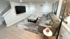 Uma sala de estar moderna com uma TV de tela plana na parede, um sofá seccional bege, uma pequena mesa de centro branca e um tapete estampado.
