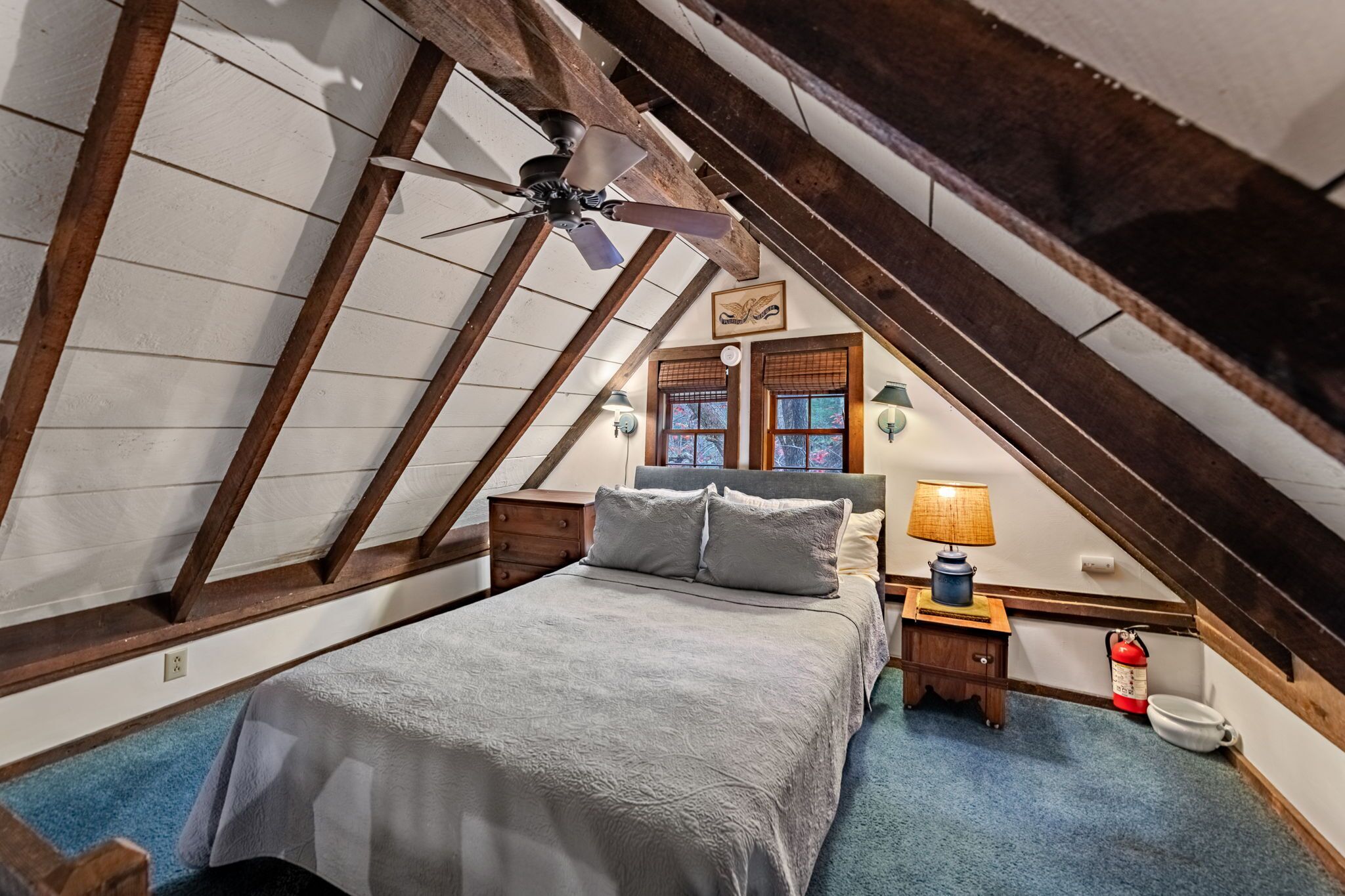 Un dormitorio con techo inclinado, vigas de madera, una cama con manta gris, una mesita de noche con lámpara y un extintor.