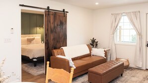 A room with a brown leather sofa, a wooden chair, a small table, a window with curtains, and a sliding door leading to another room.