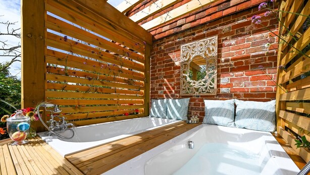 Een houten terras met twee witte whirlpools, een houten pergola en een bakstenen muur met een decoratieve spiegel.