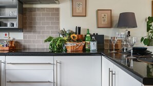 Een moderne keuken met een zwart aanrecht, witte kastjes, een roestvrijstalen afzuigkap en een achterwand van baksteen. Er hangen botanische prints in lijsten aan de muur, er staat een mand met vers fruit en een fles appelsap.