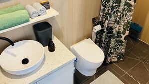Un baño con lavamanos, inodoro y cortina de ducha blancos, con un estampado de hojas. Hay toallas en un estante y un cubo de basura.