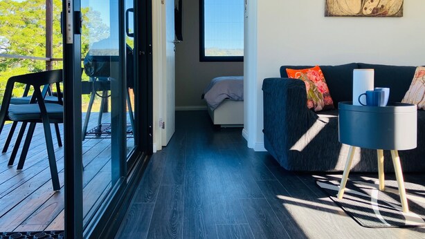 Un interior moderno con piso de madera oscura, un sofá con cojines coloridos, una mesa de centro y un ventanal que ofrece vistas al verdor.