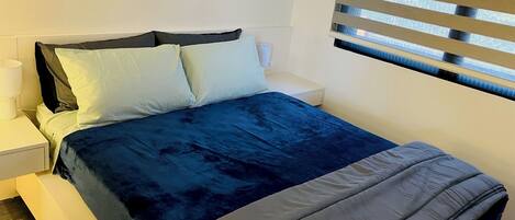 A neatly made bed with a blue and grey duvet, a white nightstand with a lamp, and a window with horizontal blinds.