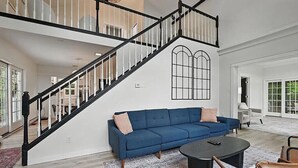 Een ruime woonkamer met een blauwe bank, een salontafel en een trap met zwarte leuningen.