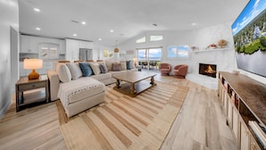 Ein geräumiges Wohnzimmer mit einer großen Couch, einem Couchtisch und einem Kamin. Es gibt eine Küchenzeile mit weißen Schränken und einem Esstisch.