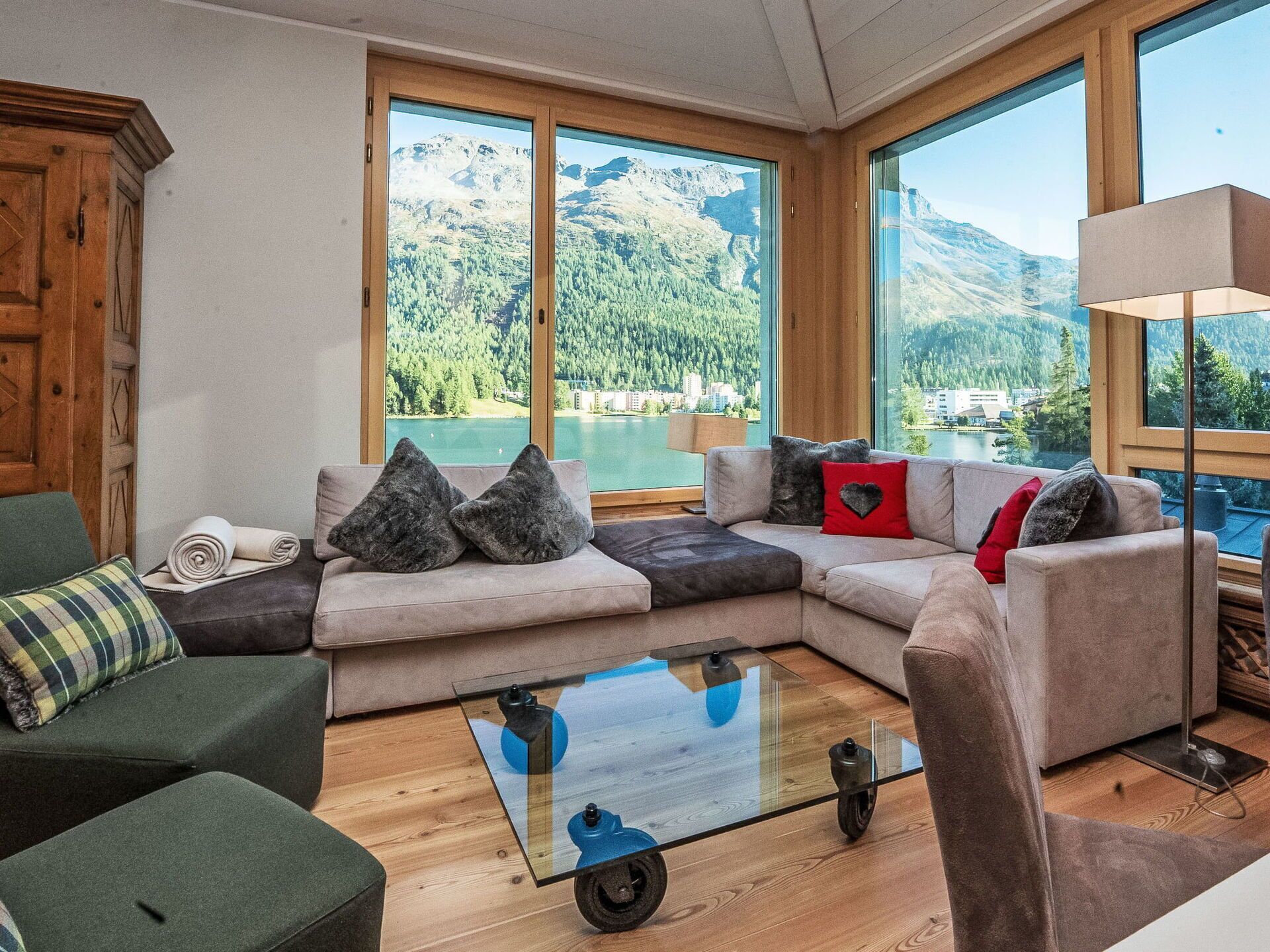 Una sala de estar moderna con un sofá grande, una mesa de centro de vidrio y vistas a un lago y montañas a través de amplios ventanales.
