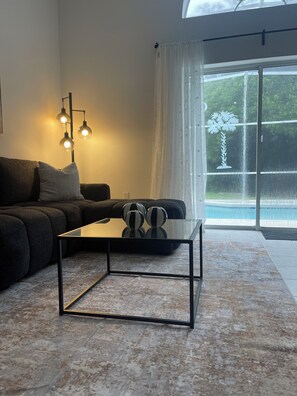 Un salon moderne avec un canapé d’angle, une table basse en verre et une vue sur une piscine, visible à travers une porte vitrée.