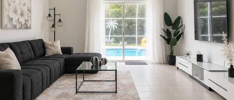 A spacious living room with a black sofa, a glass coffee table, and a large window with a view of a swimming pool.