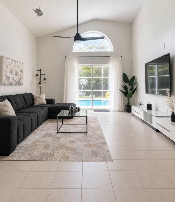 A spacious living room with a black sofa, a glass coffee table, and a large window with a view of a swimming pool.