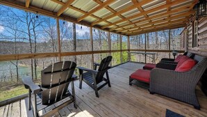 A covered deck with wooden flooring, a wicker sofa with red cushions, and two Adirondack chairs.