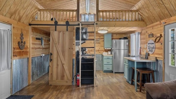 A cabin interior with a loft bedroom, a kitchen with a refrigerator and sink, and a dining area with a table and chairs.