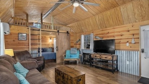 A cozy loft-style living room with a wooden ceiling, a ceiling fan, a large flat-screen TV, a sofa, a blue armchair, and a wooden coffee table.
