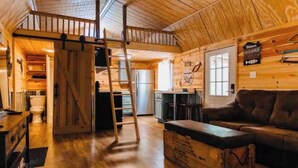 A rustic cabin interior with a wooden floor, a loft area, a kitchen, and a living room with a couch.