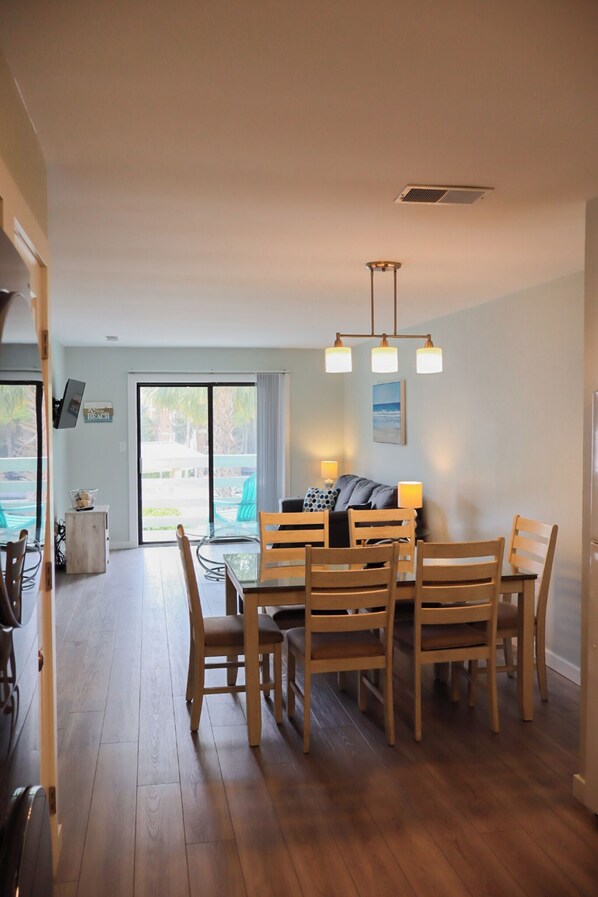 A dining area with a wooden table and chairs, a ceiling light fixture, and a view through a glass door to an outdoor area with chairs.