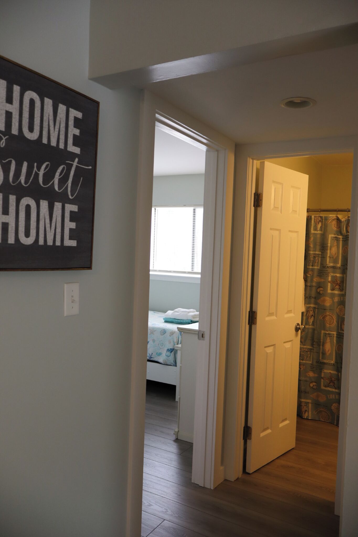 A hallway with a sign that reads "HOME is sweet HOME," leading to a bedroom with a bed and a window.