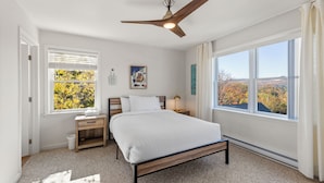 A bedroom with a bed, a nightstand, a desk, a window with blinds, a ceiling fan, and a view of autumn foliage. - Amazing Mountain Views  (Lake Placid)