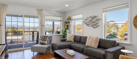 Un salón amplio con un sofá seccional gris, una mesa de centro de madera y vistas al exterior a través de amplias ventanas.