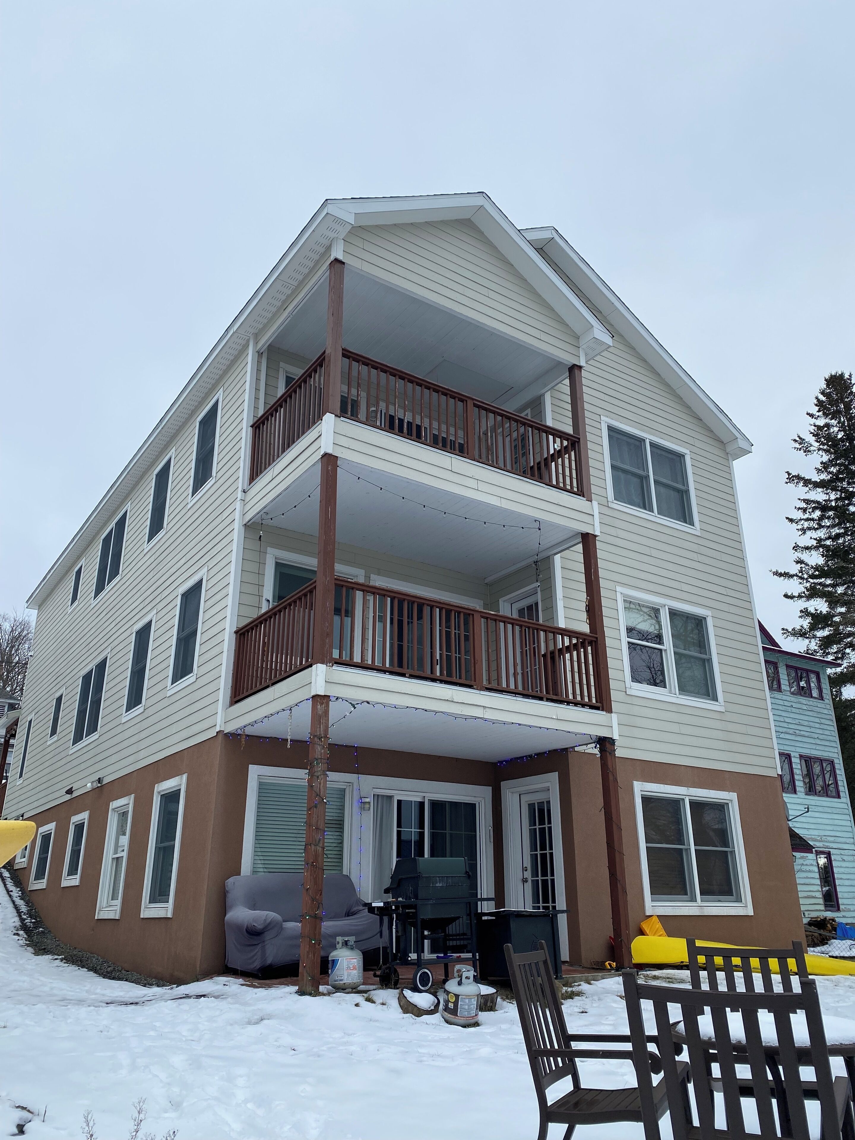 A multi-story residential building with a balcony on the second floor, a covered porch area, and a front yard with a bench and outdoor furniture.
