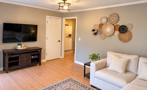 A living room with a flat-screen TV, a dark wooden entertainment unit, a light-colored sofa, a patterned rug, and a wall with woven wall art. - Adventure Hub/Near Pisgah National Forest, SierraNevada, Restaurants & Breweries (Mills River)