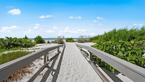 Un sentier sablonneux qui mène à une plage, avec un ciel bleu dégagé et une vue sur l’océan à l’horizon.