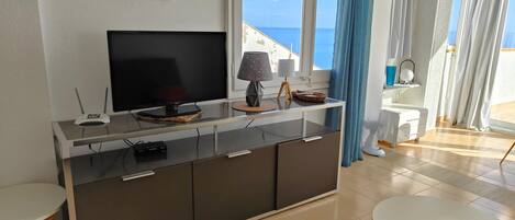 A modern living room with a flat-screen TV, a small table, a sofa, and a view of the sea through large windows.